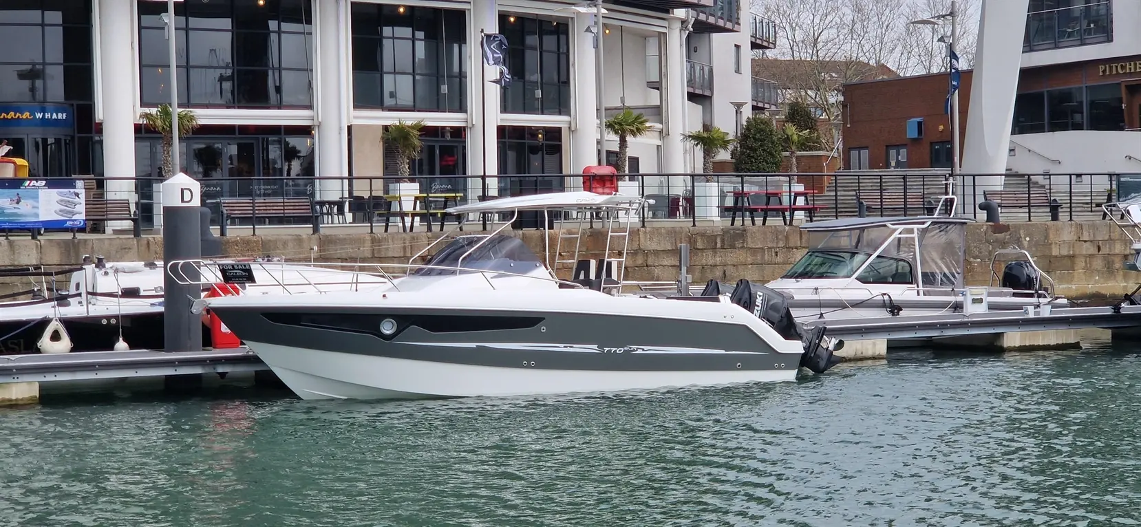 A sleek motorboat docked at a marina near a modern waterfront building with palm trees and seating areas.